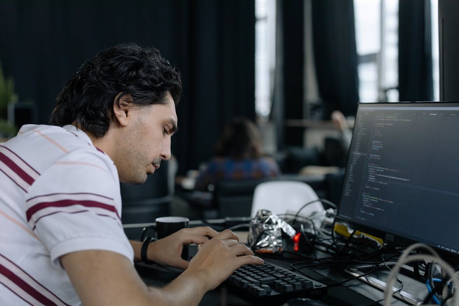 A young man with dark, wavy hair and light skin, wearing a white polo shirt with maroon stripes, is seated at a desk in an office environment during daytime. He is focused on a keyboard and mouse, wor